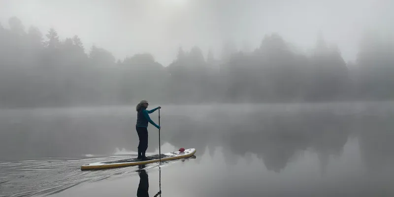 Femme debout sur un paddle, rame à la main, lac calme au lever du jour